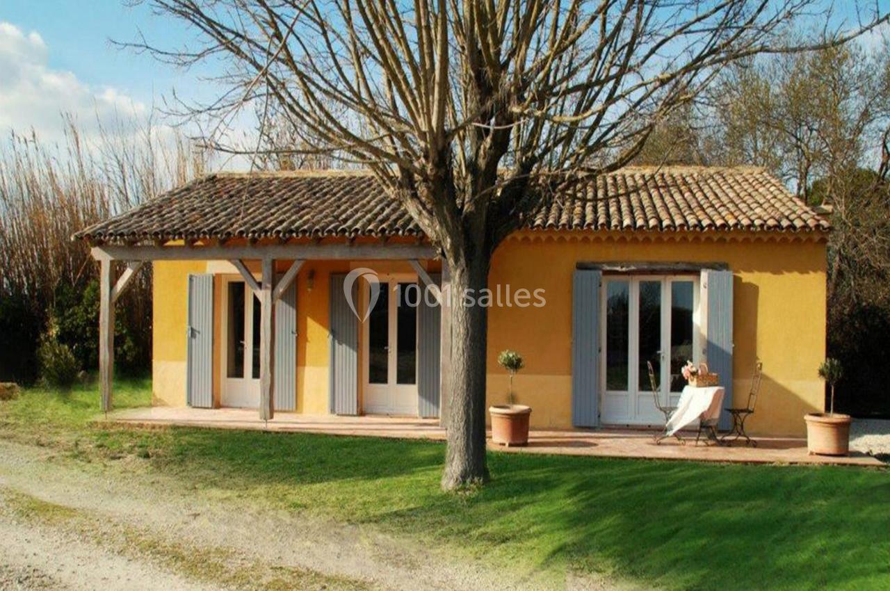 Maison de plain-pied jaune avec volets bleus, terrasse couverte, arbre au premier plan et jardin verdoyant.