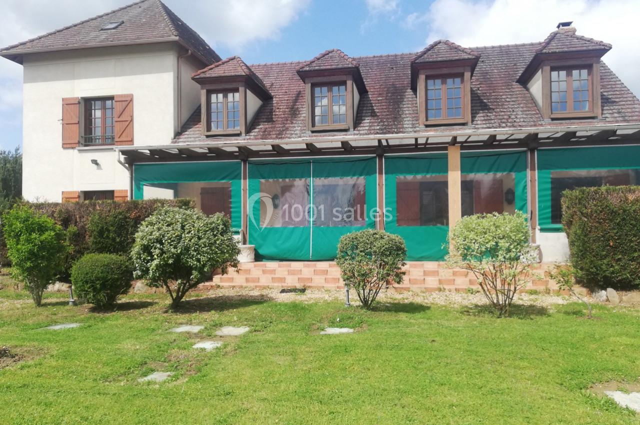 Façade d'une maison avec toit en tuiles, volets en bois, terrasse couverte et jardin arboré au premier plan.