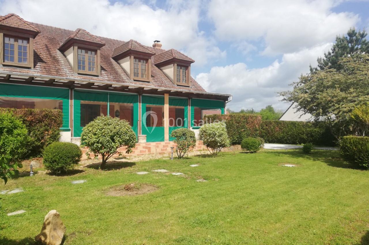 Maison en pierre avec toit en tuiles, entourée d'un jardin verdoyant et d'arbustes sous un ciel partiellement nuageux.