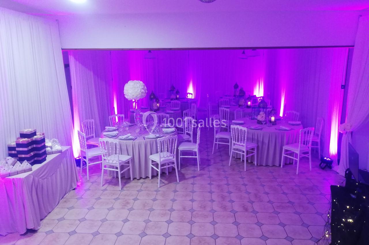 Salle de réception décorée avec des tables rondes, nappes grises, chaises blanches et éclairage violet.