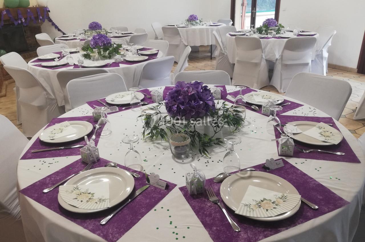 Salle décorée pour un événement avec des tables rondes dressées, nappes blanches et accents violets.
