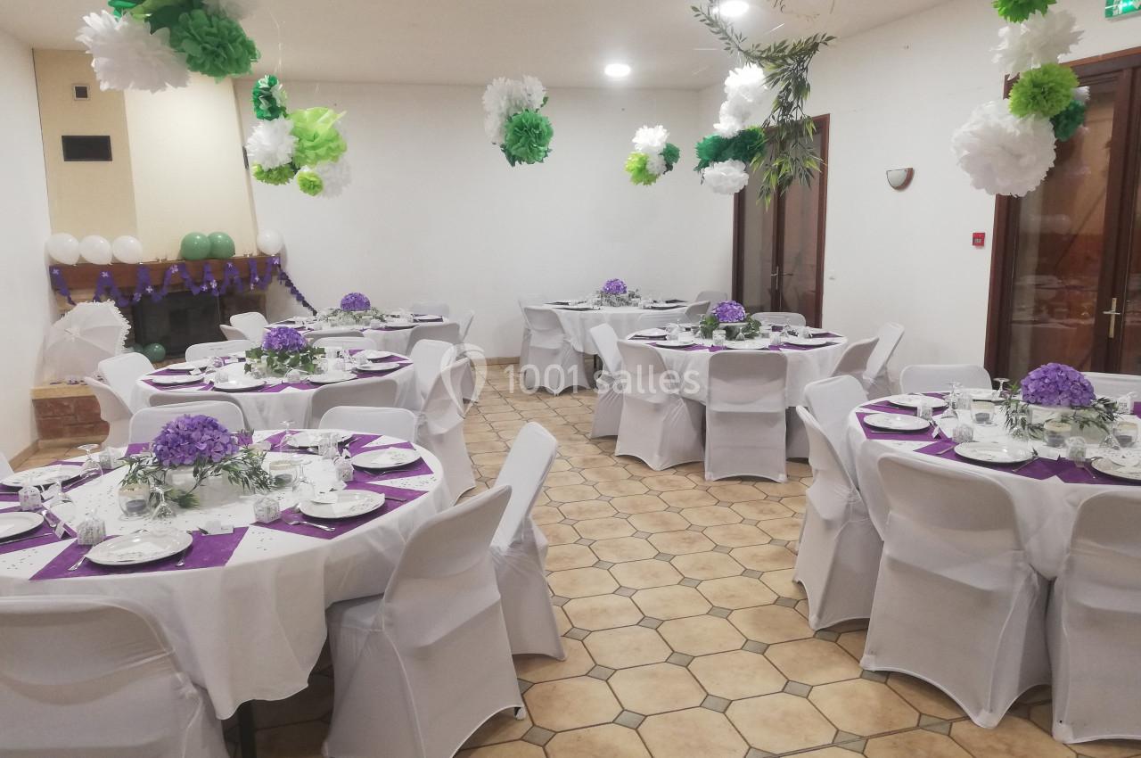 Salle décorée pour un événement avec des tables rondes dressées, nappes blanches, centres de table violets et guirlandes…