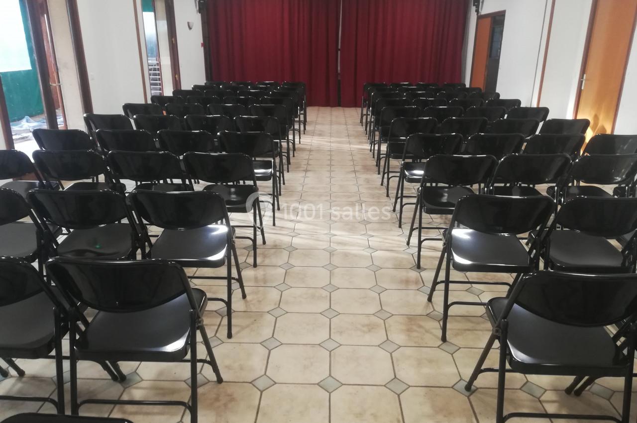 Salle avec des rangées de chaises noires pliantes alignées face à une scène avec un rideau rouge.
