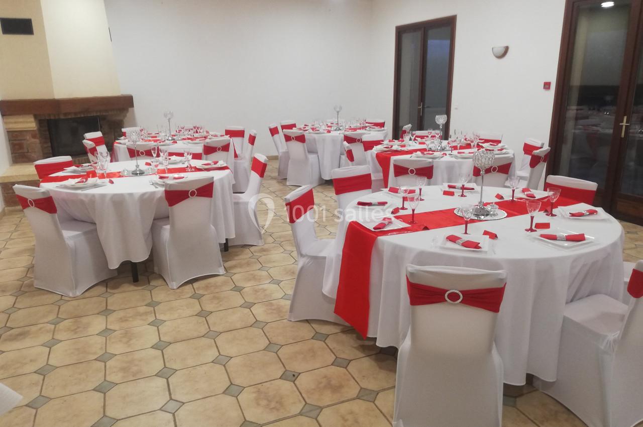Salle de réception avec tables rondes dressées, nappes blanches et décorations rouges, sol en carrelage beige.