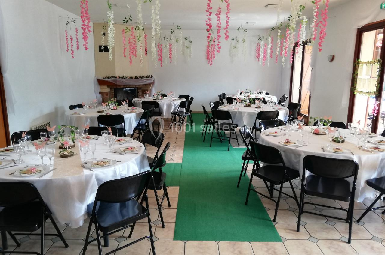 Salle décorée pour un événement avec des tables rondes dressées, nappes blanches et guirlandes florales suspendues.