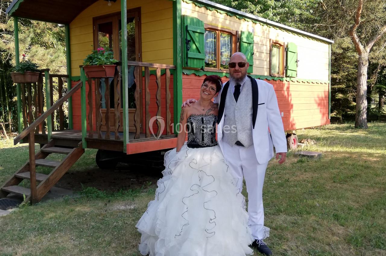Un couple en tenue de mariage pose devant une roulotte colorée dans un cadre verdoyant.