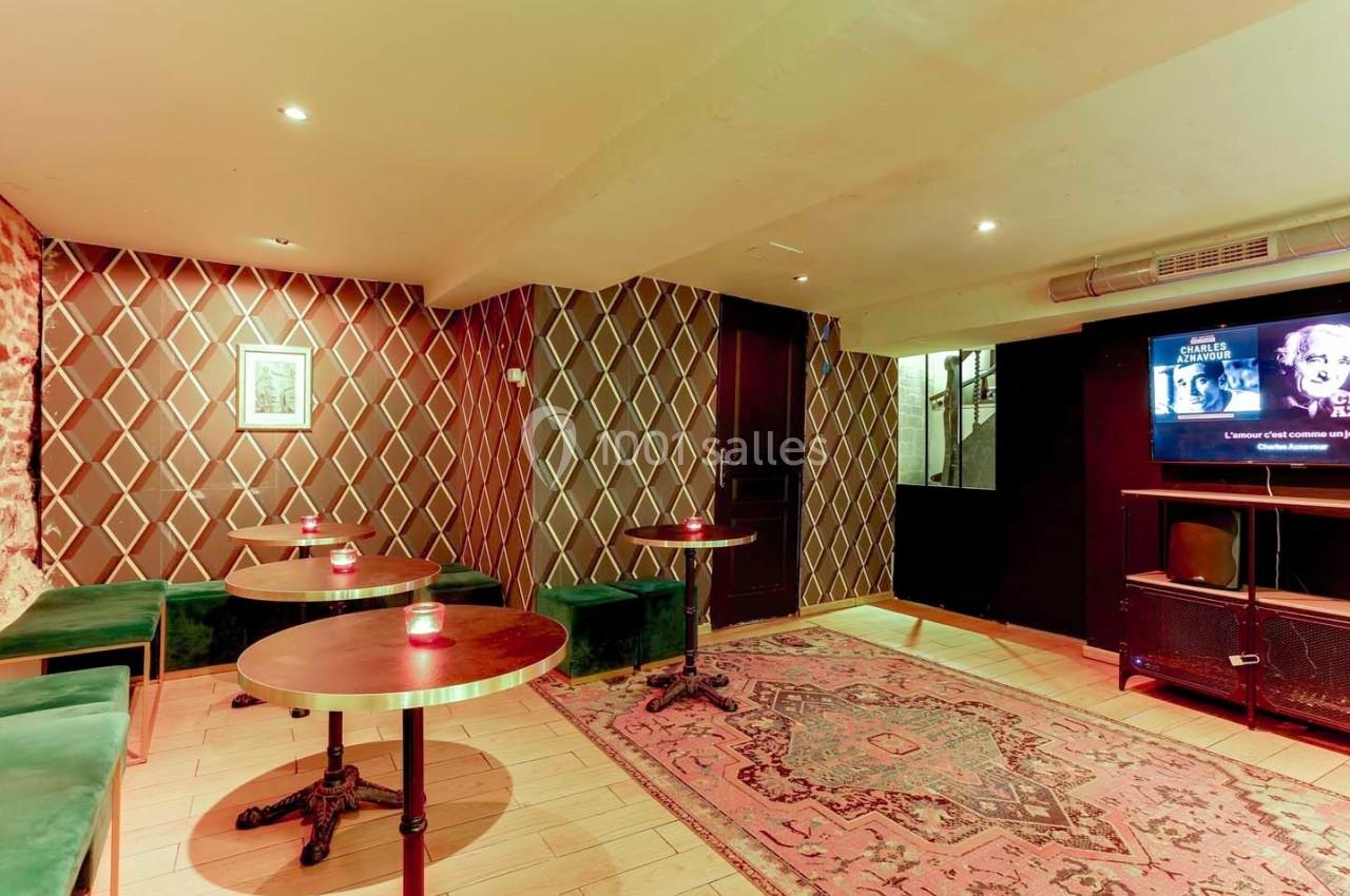 Salle avec tables rondes, banquettes vertes, tapis coloré et écran affichant une image, éclairée par des lumières tamisées.