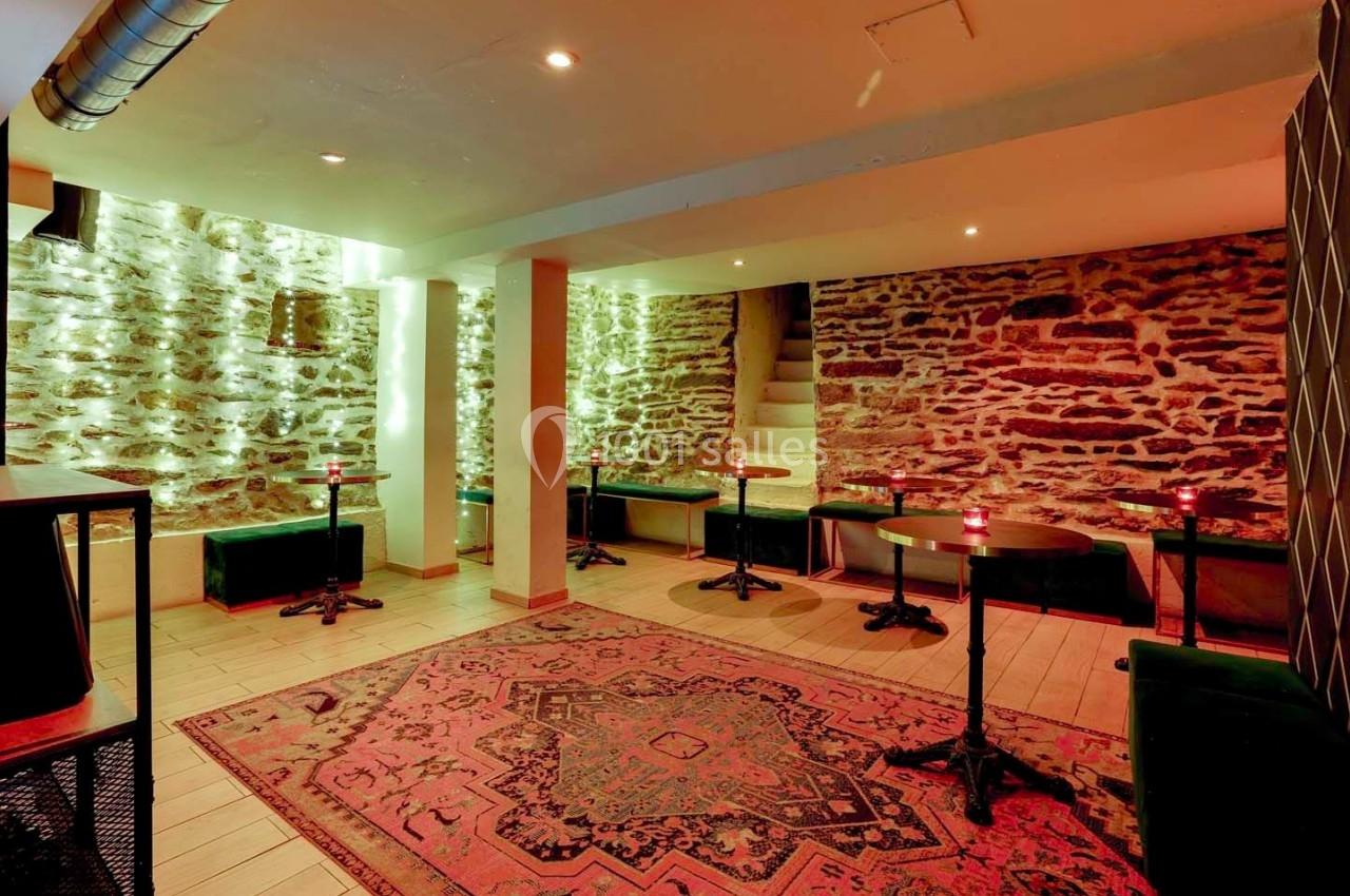 Salle voûtée avec murs en pierre apparente, tables rondes, banquettes vertes et tapis coloré au sol.