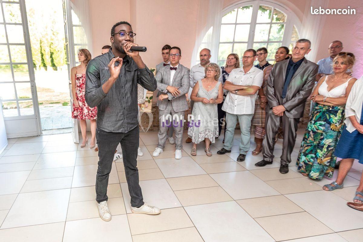 Un homme chante au micro devant un groupe de personnes debout dans une salle lumineuse avec de grandes fenêtres.