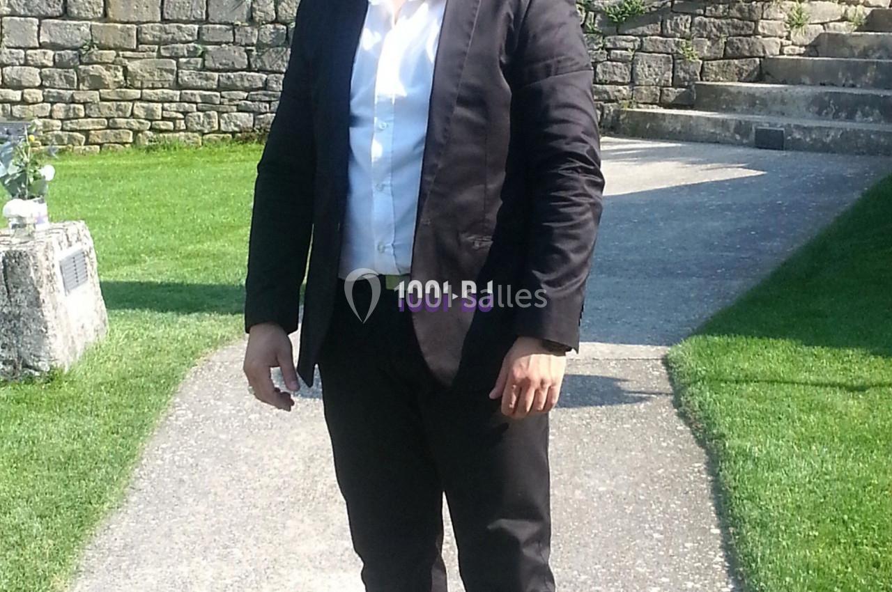 Homme en costume noir debout sur une allée de jardin, entouré de pelouse et de parterres de fleurs.