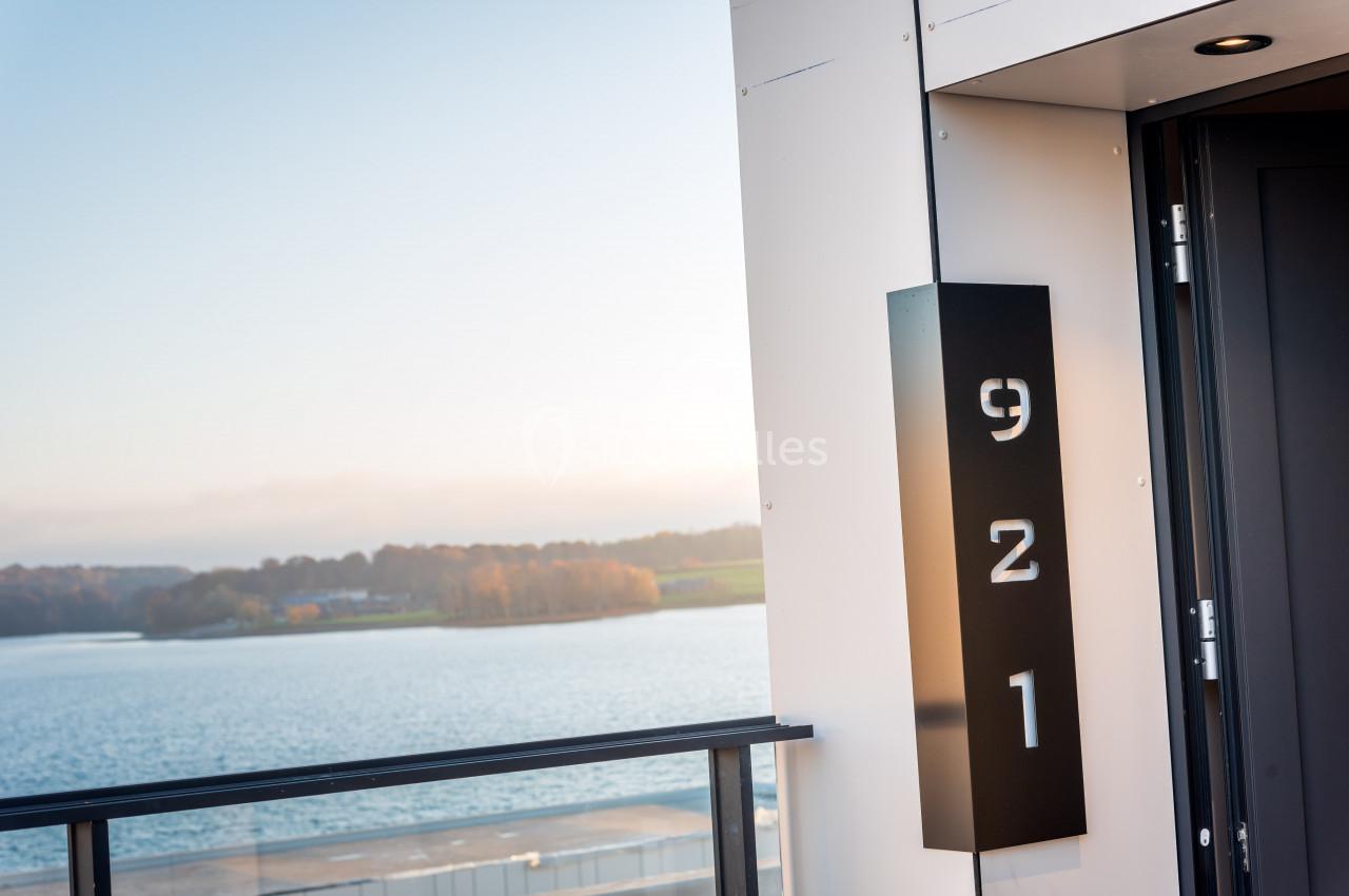 Plaque noire avec le numéro ’921’ fixée sur un mur, vue sur un lac et un paysage boisé en arrière-plan.