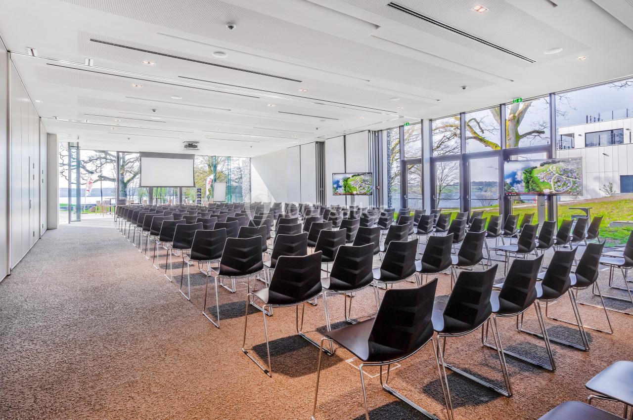 Salle de conférence lumineuse avec rangées de chaises noires, écrans et grandes baies vitrées donnant sur un espace…