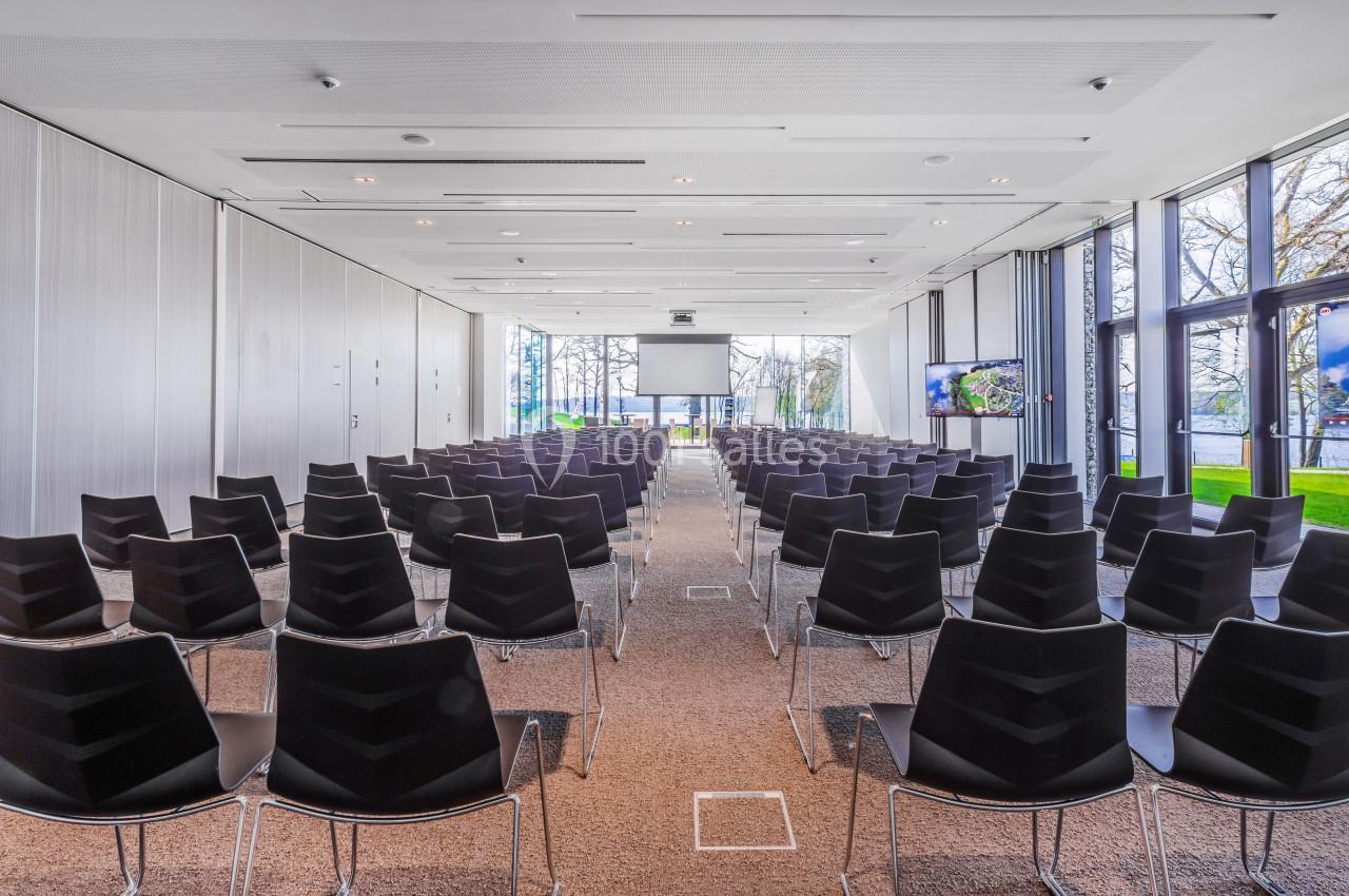 Salle de conférence lumineuse avec des rangées de chaises noires alignées face à un écran de projection.