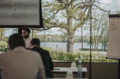 Terrasse en bois avec table et chaises, vue sur un jardin verdoyant et un lac au coucher du soleil.