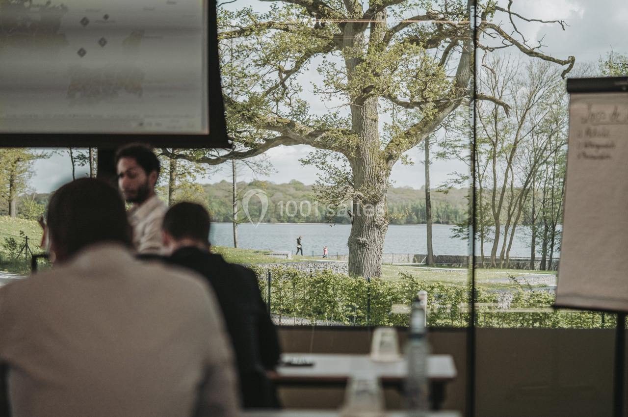 Vue depuis une salle de réunion avec des participants, donnant sur un lac entouré d'arbres et un chemin piéton.