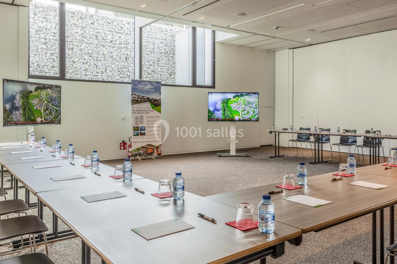 Salle de réunion lumineuse avec tables en U, chaises, écrans, bouteilles d'eau et documents disposés sur les tables.