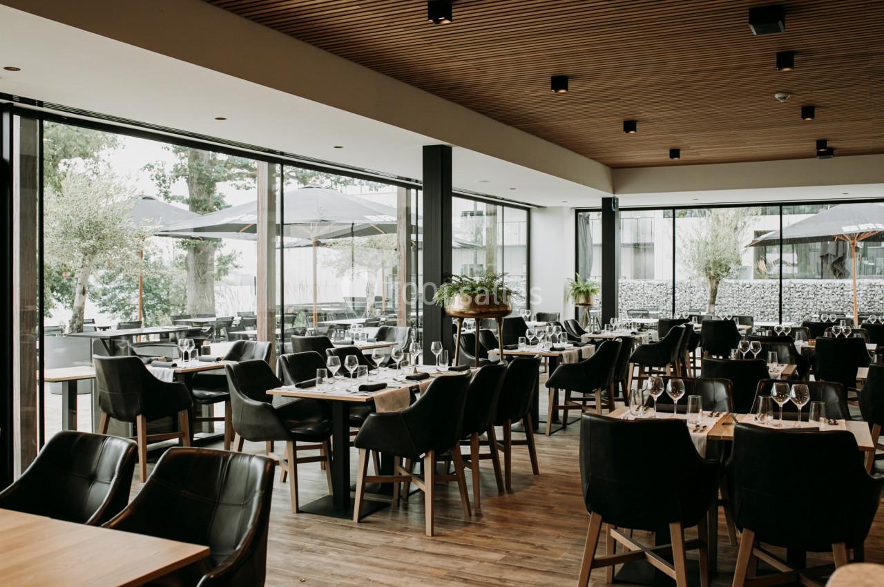 Salle de restaurant moderne avec tables dressées, chaises noires et grandes baies vitrées donnant sur une terrasse.