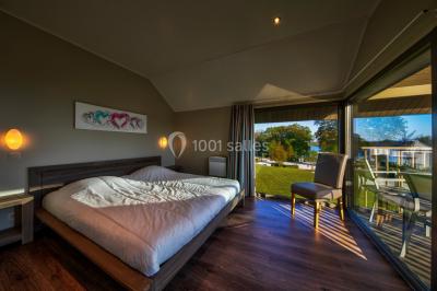 Terrasse en bois avec table et chaises, vue sur un jardin verdoyant et un lac au coucher du soleil.