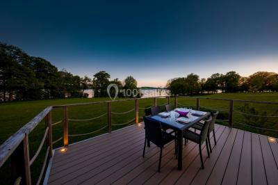 Terrasse en bois avec table et chaises, vue sur un jardin verdoyant et un lac au coucher du soleil.