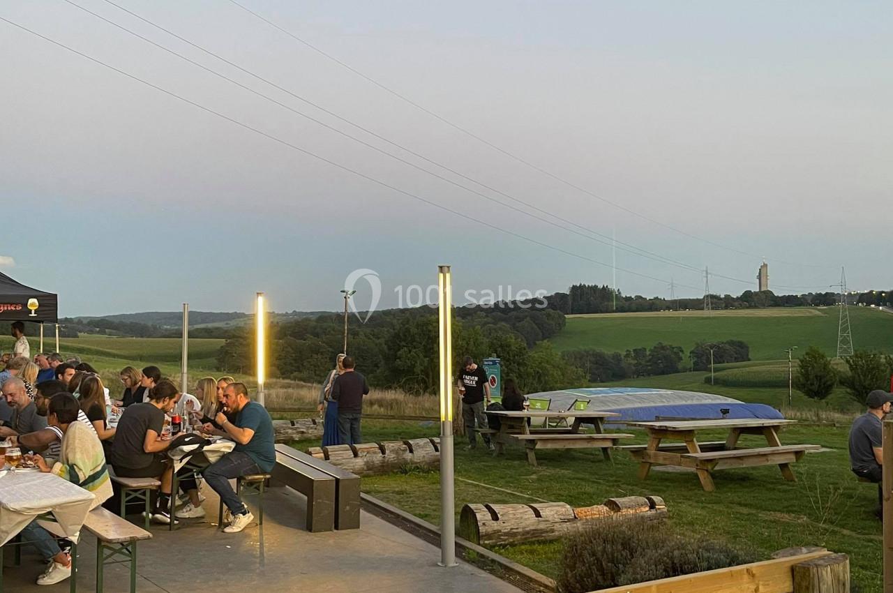 Des personnes dînent en plein air sur des tables en bois, avec une vue sur des collines et un ciel dégagé en arrière-plan.