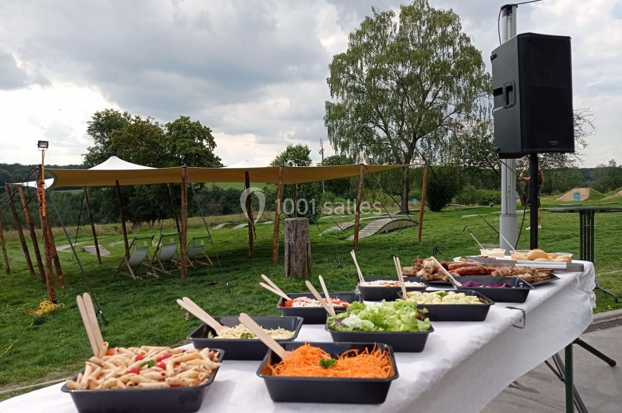 Buffet en plein air avec divers plats et salades sur une table, dans un cadre verdoyant sous un ciel nuageux.