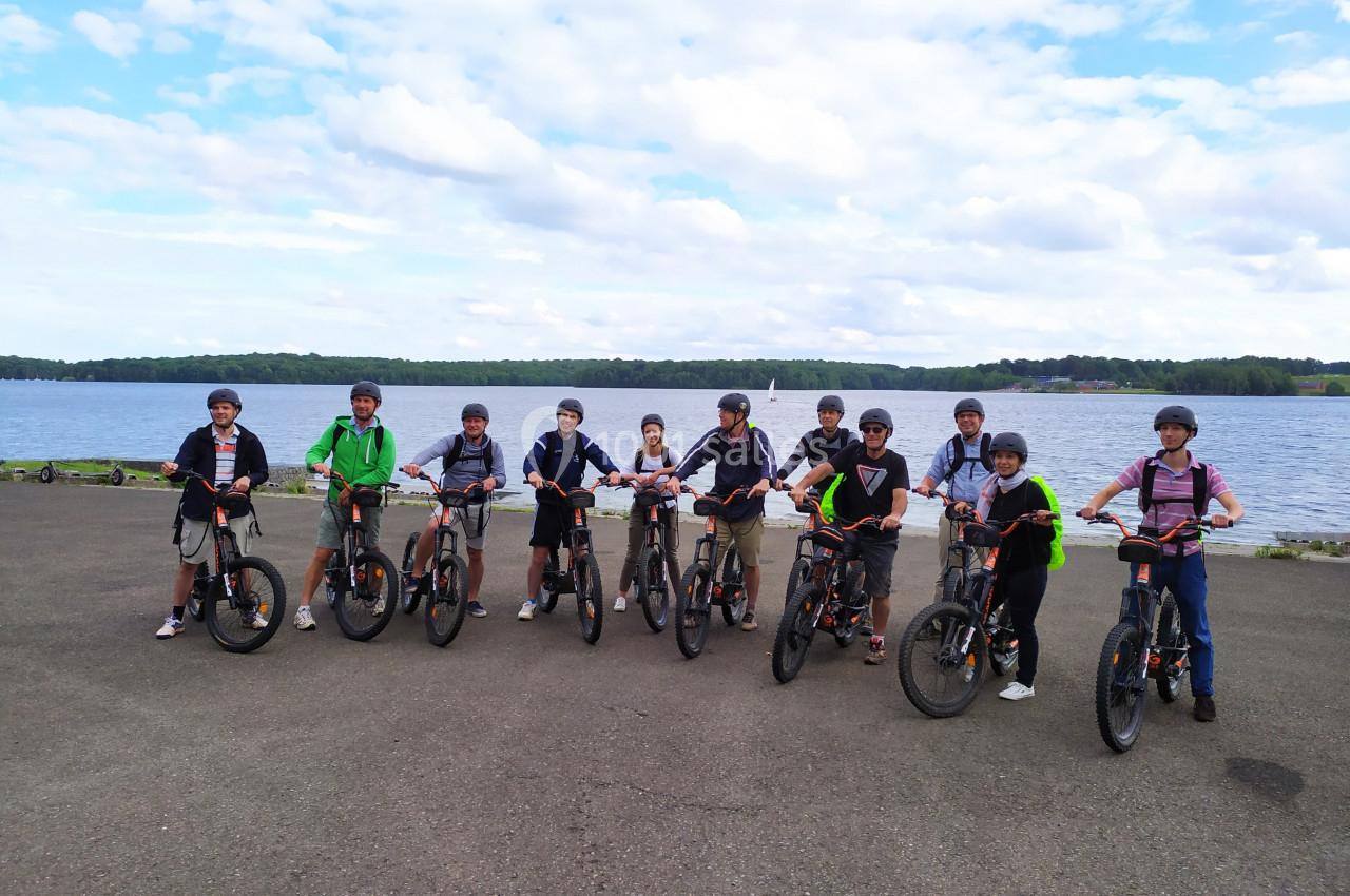 Un groupe de personnes en tenue décontractée posant avec des vélos devant un lac sous un ciel partiellement nuageux.