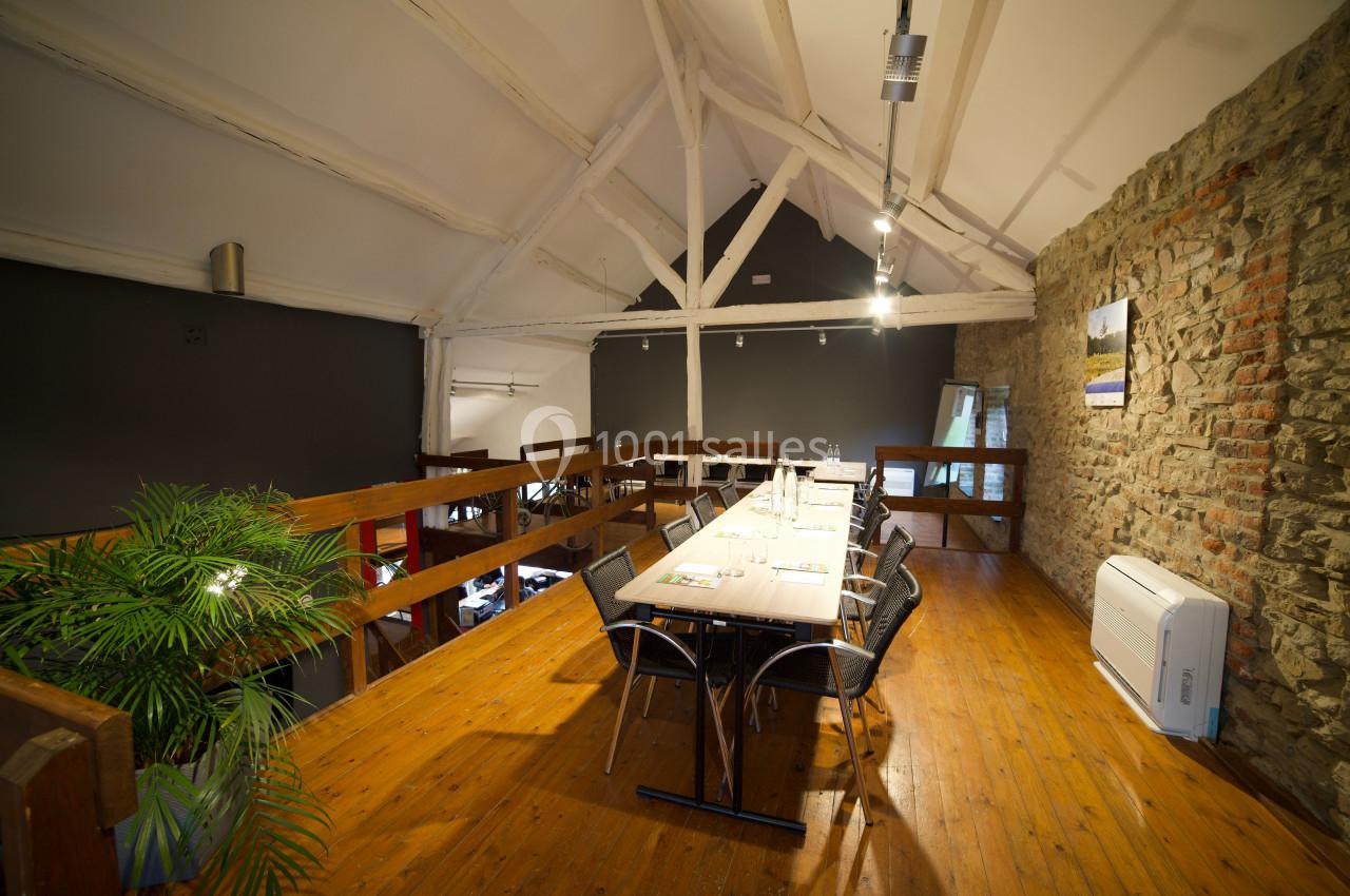 Salle de réunion en mezzanine avec table en bois, chaises noires, mur en pierre et plafond avec poutres apparentes.