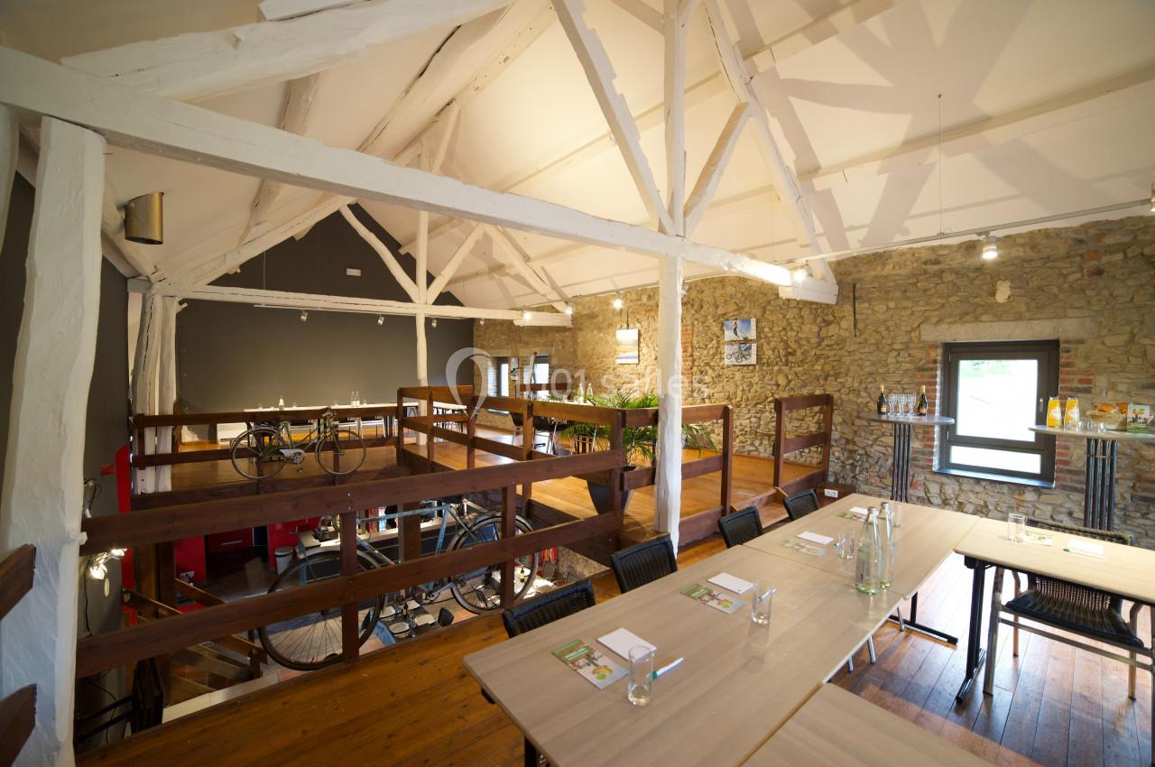 Salle lumineuse avec poutres apparentes, murs en pierre, tables dressées et mezzanine avec des vélos exposés.