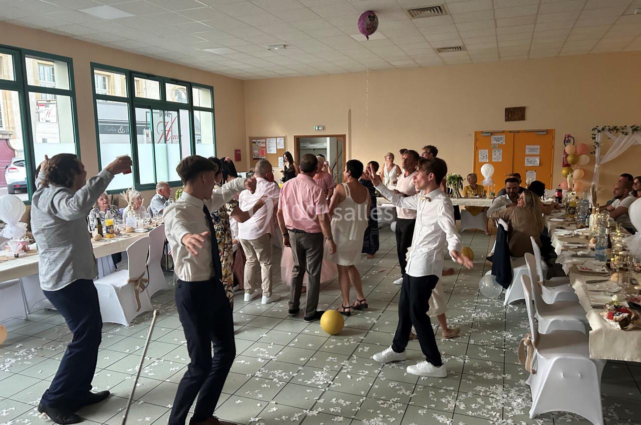 Des invités dansent dans une salle décorée pour une réception, avec des tables dressées et des ballons au sol.