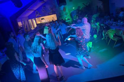 Des personnes dansent dans une salle éclairée par des lumières rouges, entourées de tables dressées pour un événement.