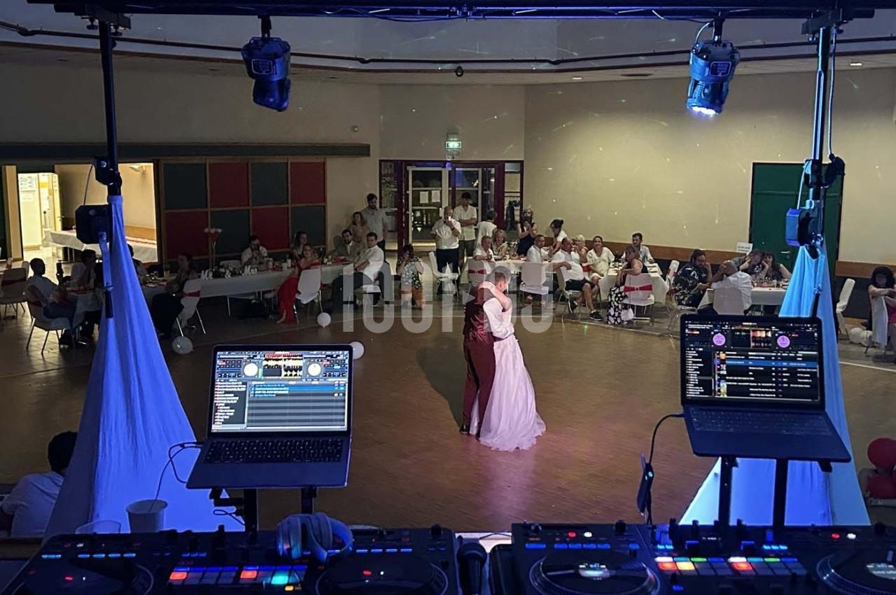Un couple danse au centre d'une salle de réception, vu depuis une table de DJ équipée de matériel audio.
