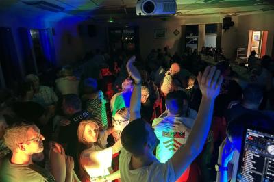 Des personnes dansent dans une salle éclairée par des lumières rouges, entourées de tables dressées pour un événement.