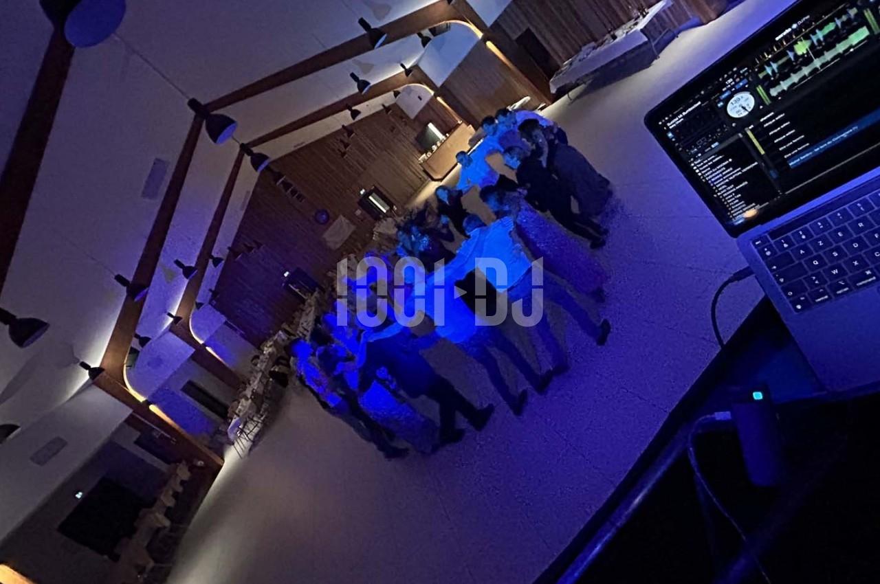 Groupe de personnes dansant en cercle dans une salle éclairée par des lumières bleues, avec un ordinateur de DJ au premier…
