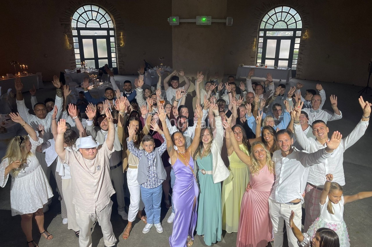 Groupe de personnes souriantes levant les bras dans une salle avec des murs en pierre et des fenêtres cintrées.