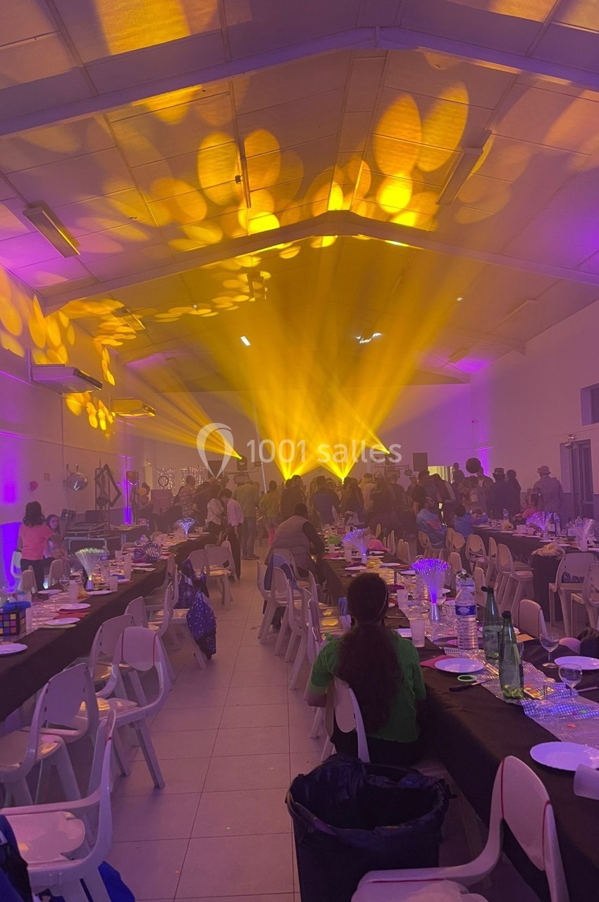 Salle de réception animée avec des invités assis, éclairage jaune vif et décorations sur les tables.