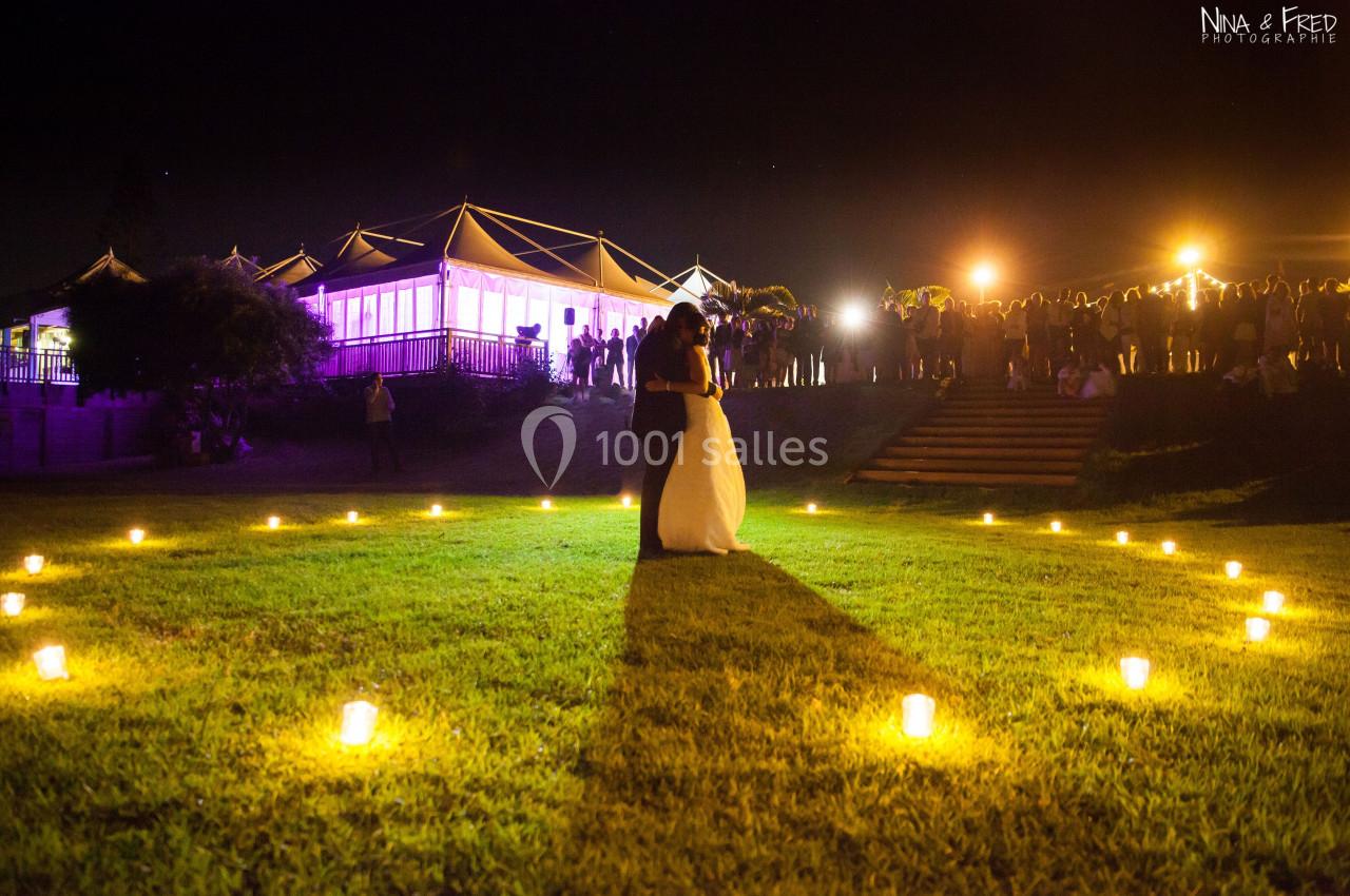 Un couple danse au centre d'une pelouse éclairée par des bougies, avec un bâtiment illuminé et des invités en arrière-plan.