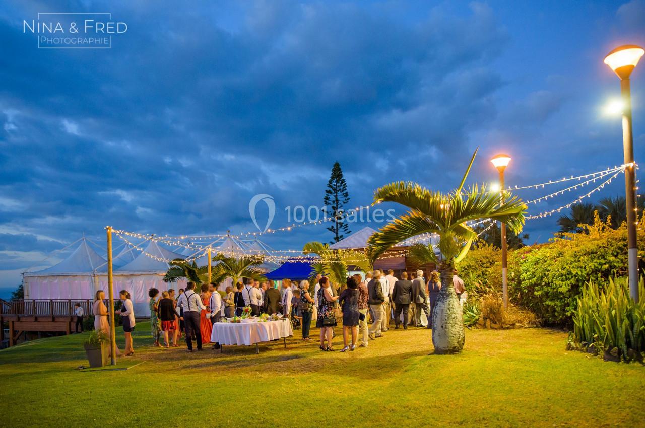 Groupe de personnes rassemblées en extérieur au crépuscule, près de tables décorées sous des guirlandes lumineuses.