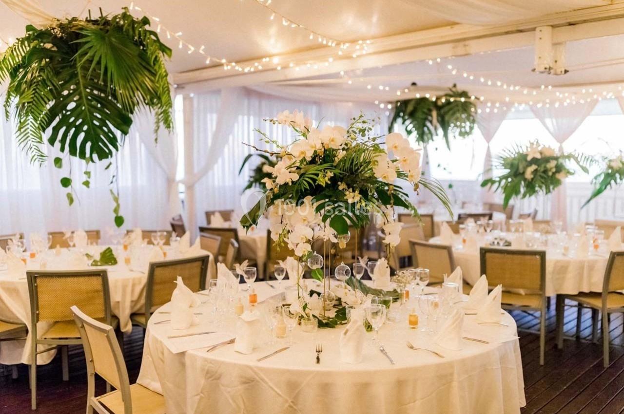Salle de réception décorée avec des tables rondes élégantes, des fleurs blanches et des plantes suspendues.