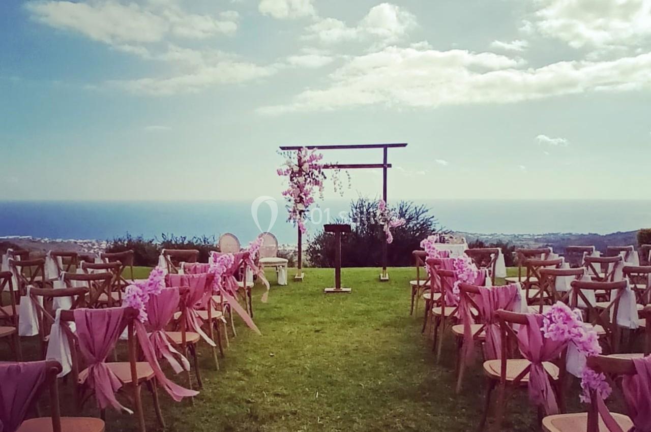 Allée décorée de chaises roses menant à une arche fleurie en extérieur, avec vue sur la mer et le ciel.