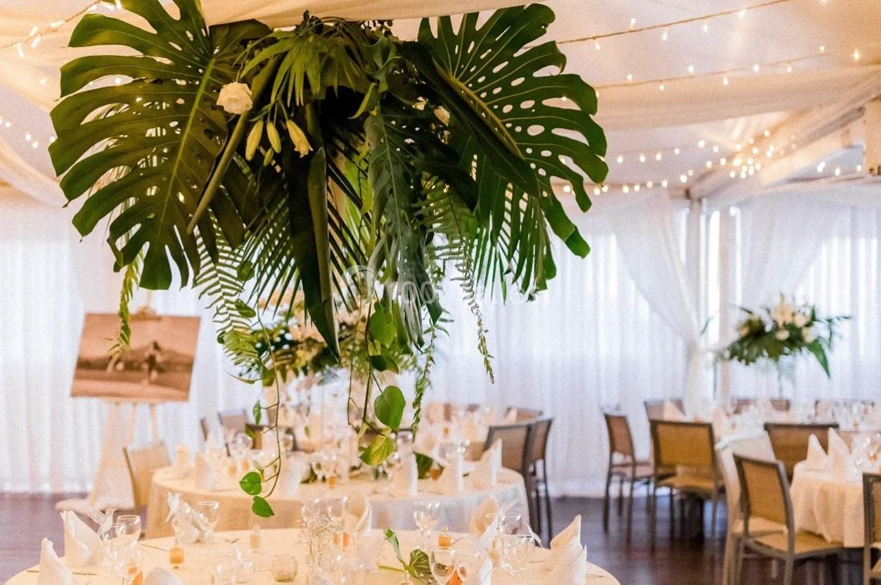 Salle de réception décorée avec des tables dressées, des nappes blanches et une suspension végétale au centre.