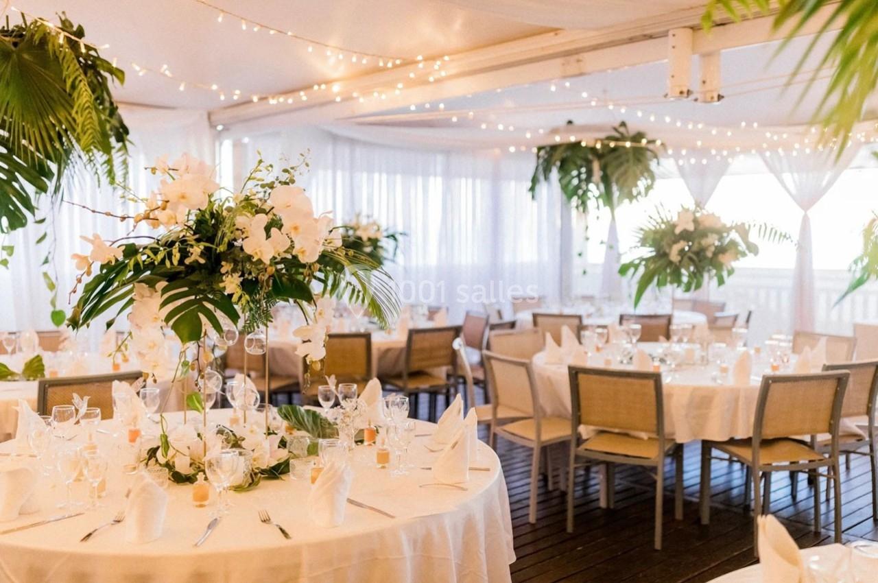Salle de réception décorée avec des tables rondes, nappes blanches, compositions florales et guirlandes lumineuses…