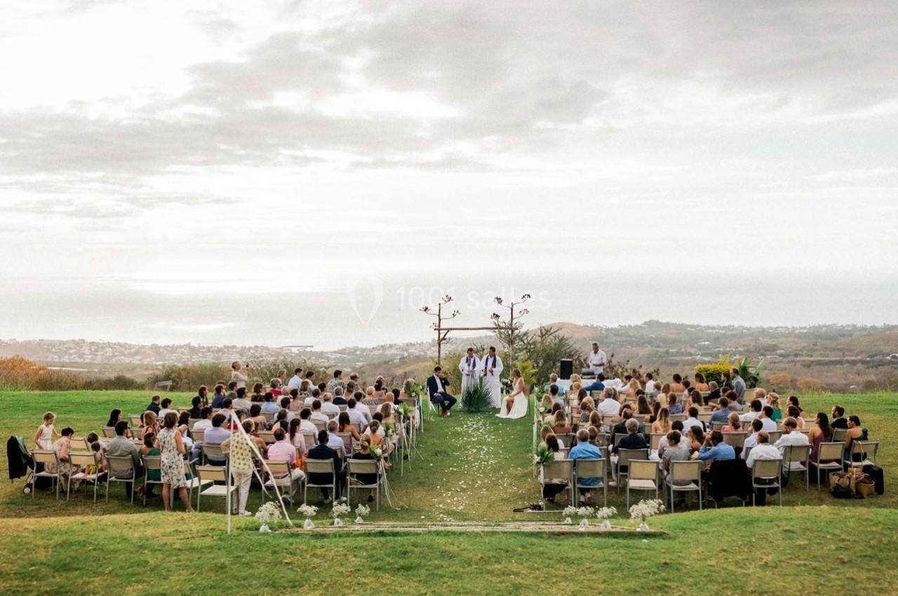 Cérémonie de mariage en plein air avec des invités assis face à un autel décoré, vue sur un paysage côtier en arrière-plan.