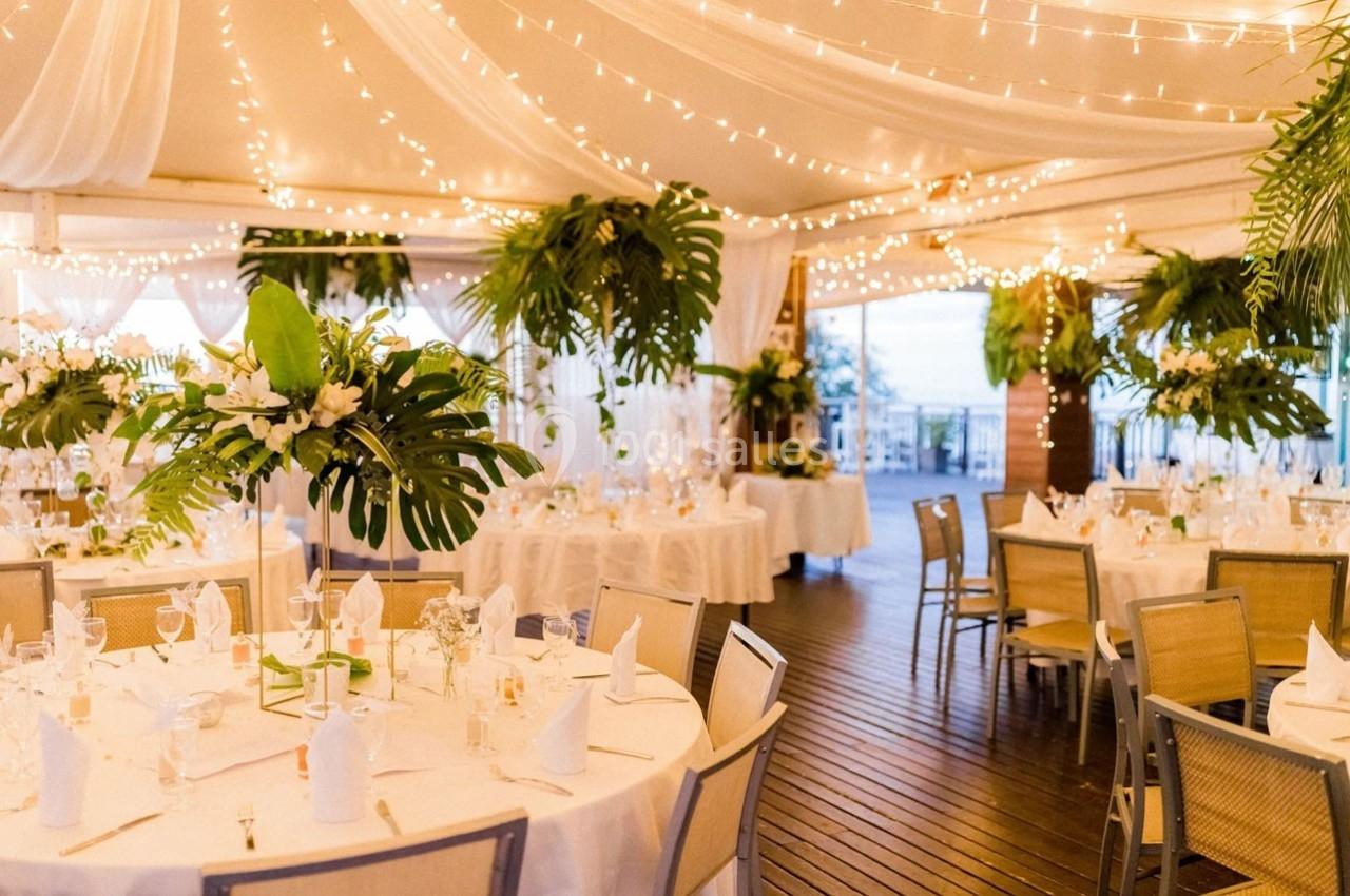 Salle de réception décorée avec des guirlandes lumineuses, des tables dressées et des compositions florales tropicales.