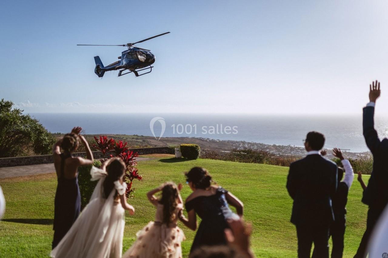 Un hélicoptère s'envole au-dessus d'un groupe de personnes sur une pelouse avec vue sur la mer.