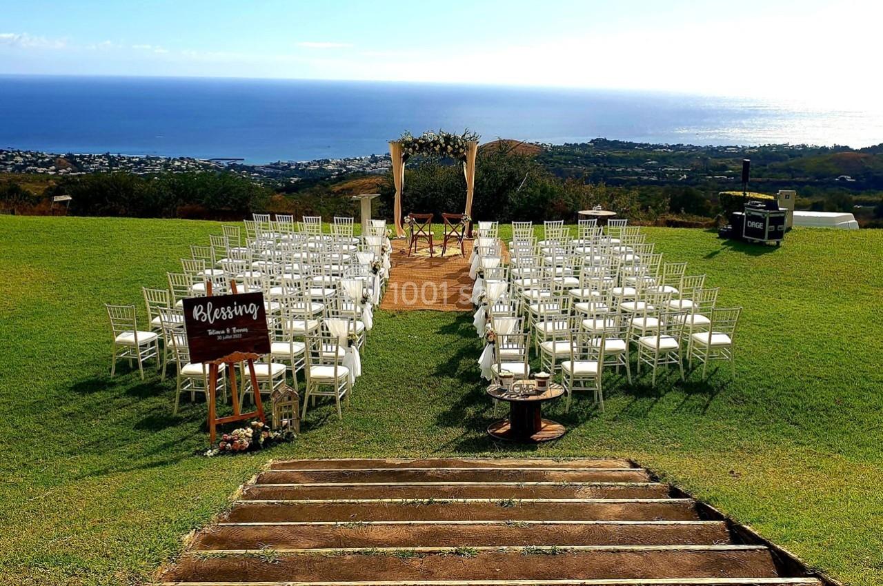 Chaises blanches disposées en rangées sur une pelouse, face à une arche décorée, avec vue sur la mer et une ville en arrière…