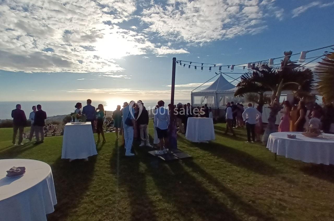 Groupe de personnes rassemblées sur une pelouse en bord de mer au coucher du soleil, avec des tables et une tente blanche.