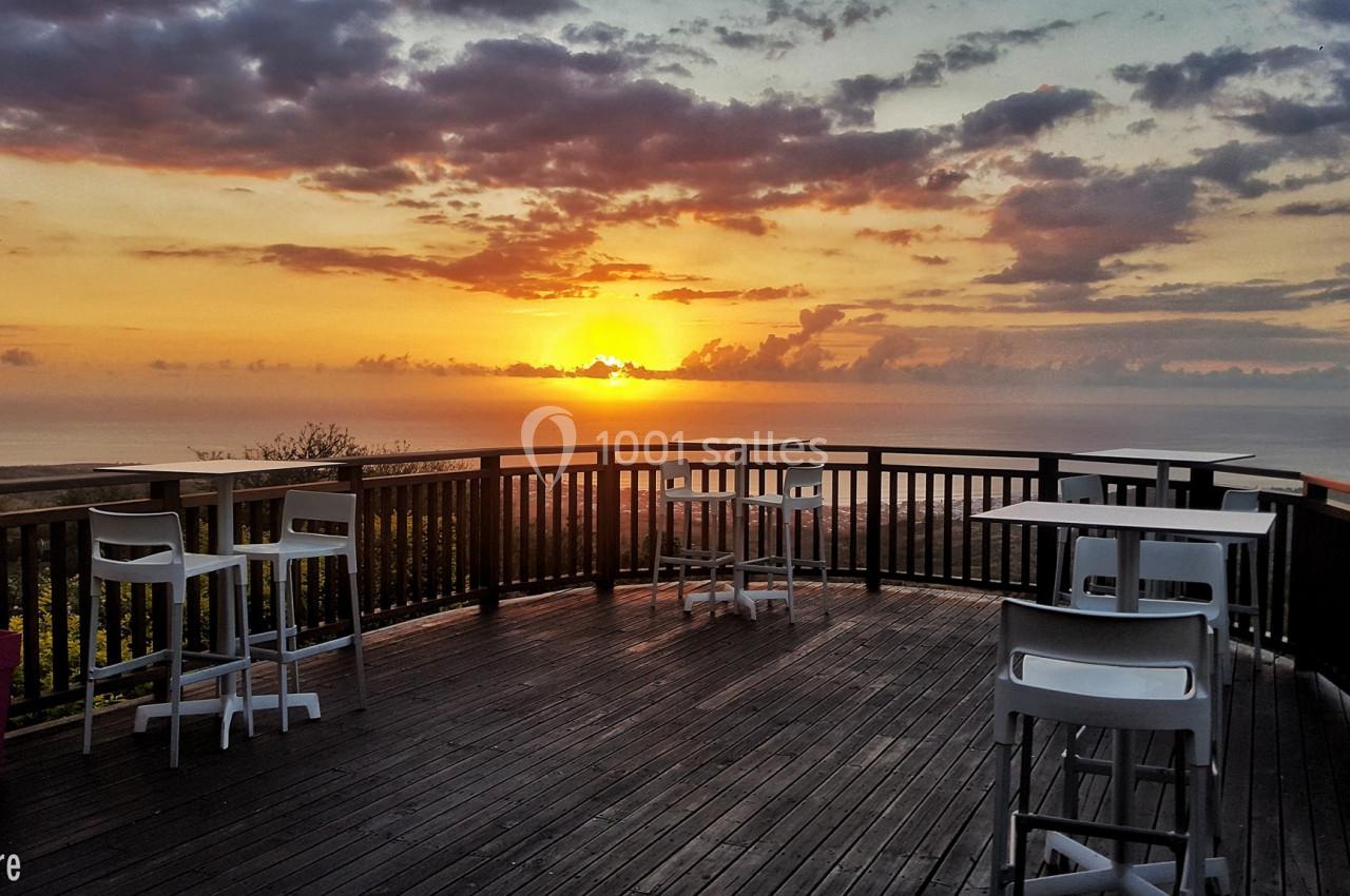 Terrasse en bois avec des tables et chaises blanches, offrant une vue sur un coucher de soleil au-dessus de l'océan.