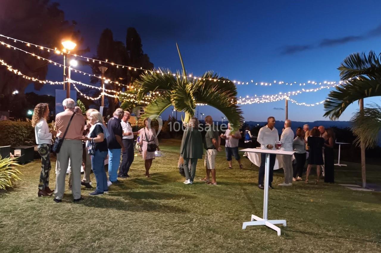 Groupe de personnes discutant en extérieur le soir, sous des guirlandes lumineuses, sur une pelouse avec vue sur la mer.