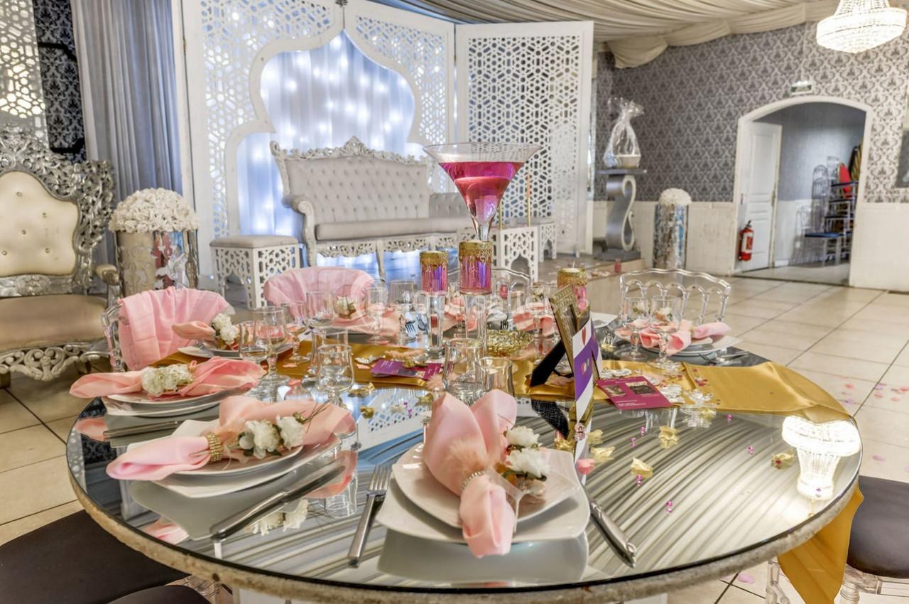 Table décorée pour un événement avec nappes roses, verres, fleurs et centre de table élégant dans une salle ornée.
