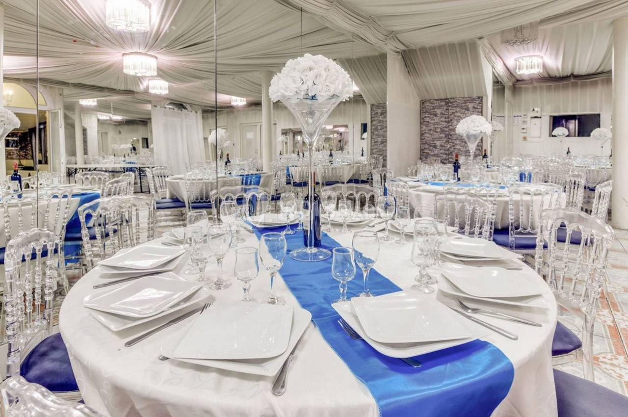 Salle de réception élégante avec tables rondes dressées, nappes blanches, chemins de table bleus et décorations florales.